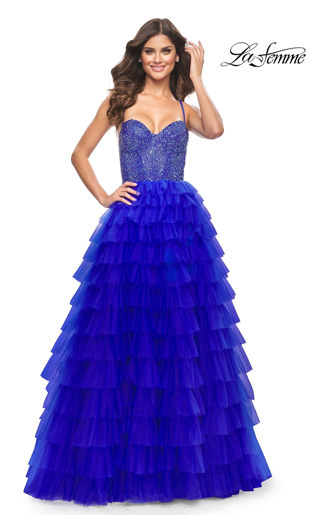 A woman smiles in the La Femme Long Ruffled Prom Ball Gown 32002—a sleeveless, floor-length blue dress with a fitted beaded bodice and tiered ruffled skirt. The logo in the top right highlights this elegant gown.